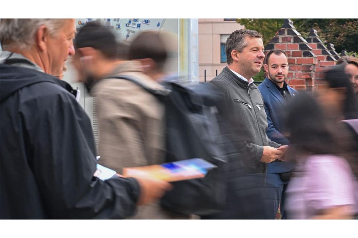 28.08.2026, Sachsen-Anhalt, Dessau-Roßlau: Sven Schulze (CDU, re. in grauer Strickjacke), Ministerpräsident von Sachsen-Anhalt und Spitzenkandidat der CDU bei der Landtagswahl, steht am 28. August in Dessau-Roßlau an einem Infostand der CDU, während Passanten auf das Gelände der Uni-Klinik eilen.