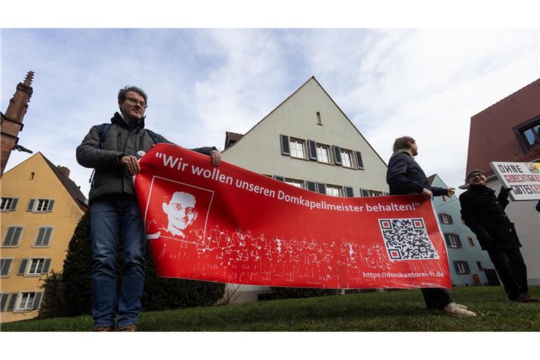 Demonstranten stehen nach einem Gottesdienst hinter dem Freiburger Münster und warten mit einem Banner mit der Aufschrift „Wir wollen unseren Domkapellmeister behalten“ (Archivbild).