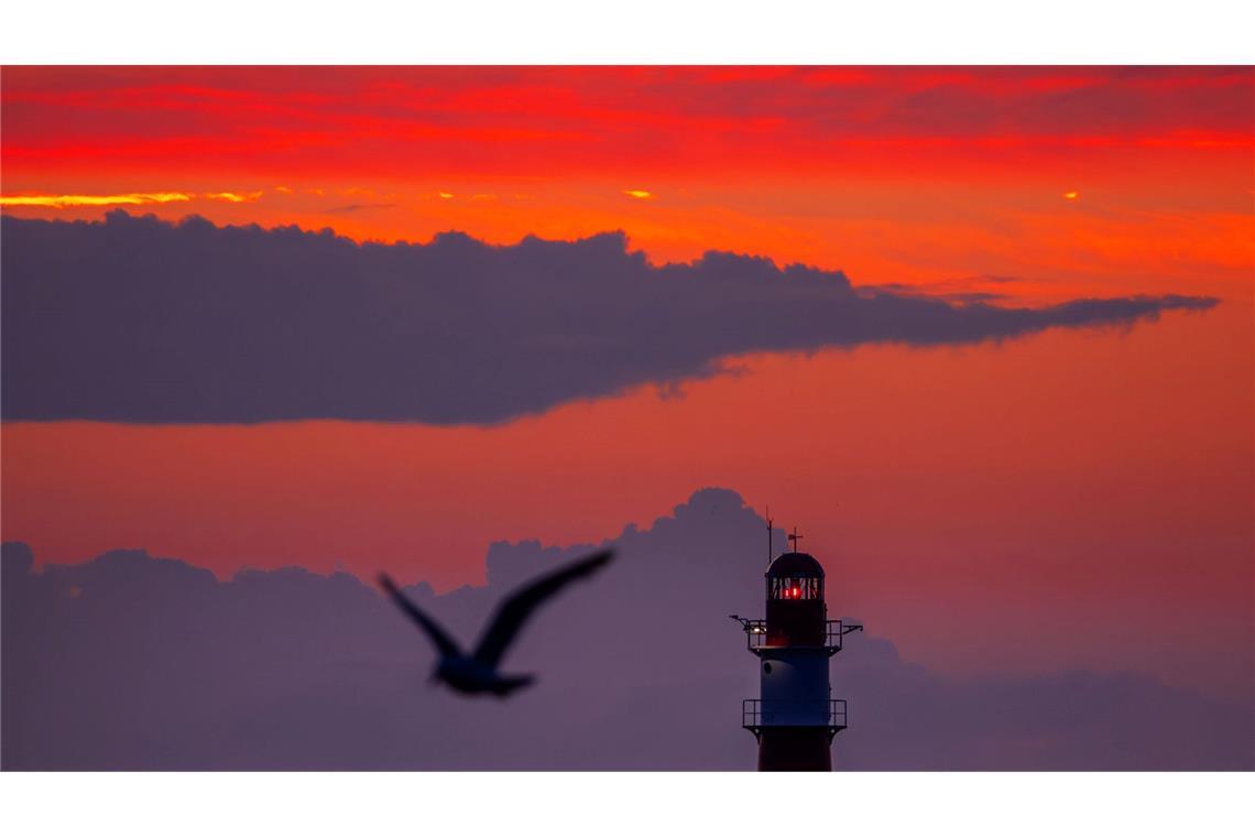 Die aufgehende Sonne färbt den Himmel hinter dem Leuchtfeuer auf der Ostmole am Ostseestrand von Warnemünde orangerot.