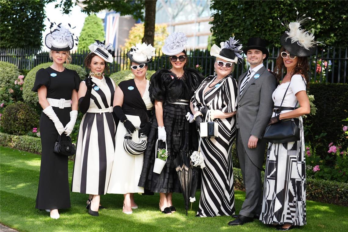 Die Gruppe setzt bei Royal Ascot auf ein konsequentes Schwarz-Weiß-Thema: gestreifte Kleider, elegante Handschuhe, Zylinder und auffällige Hüte mit Federn, Blüten und grafischen Formen ergeben einen abgestimmten, sehr klassischen Auftritt.