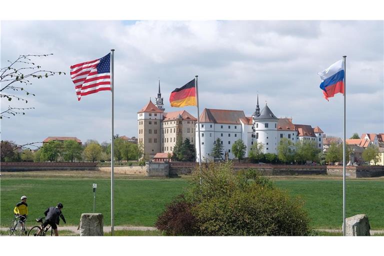 Die US-amerikanische, die deutsche und die russische Fahne wehen am Ufer der Elbe, im Hintergrund Schloss Hartenfels.