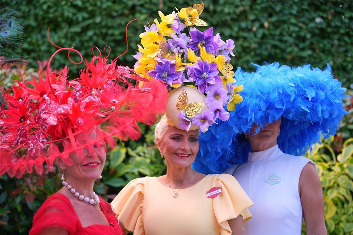 Drei Besucherinnen von Royal Ascot setzen auf maximal auffälligen Kopfschmuck: ein roter Fascinator mit Federn und floralen Elementen, ein hoher Blumenhut in Gelb und Violett sowie ein voluminöser blauer Federhut zum schlichten weißen Kleid.