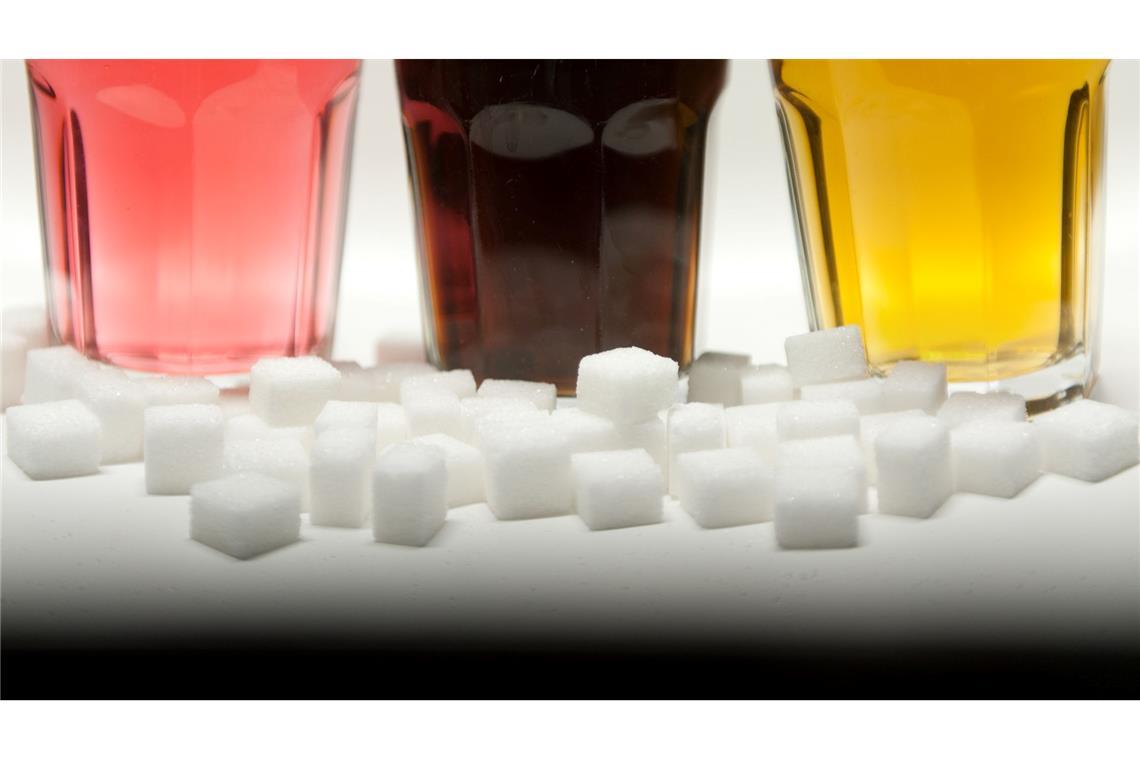Softdrinks erhöhen Risiko für Diabetes und Herzkrankheiten Drei Gläser gefüllt mit roter Limonade, Cola und einem Energy-Drink neben Zuckerwürfeln auf einem Tisch