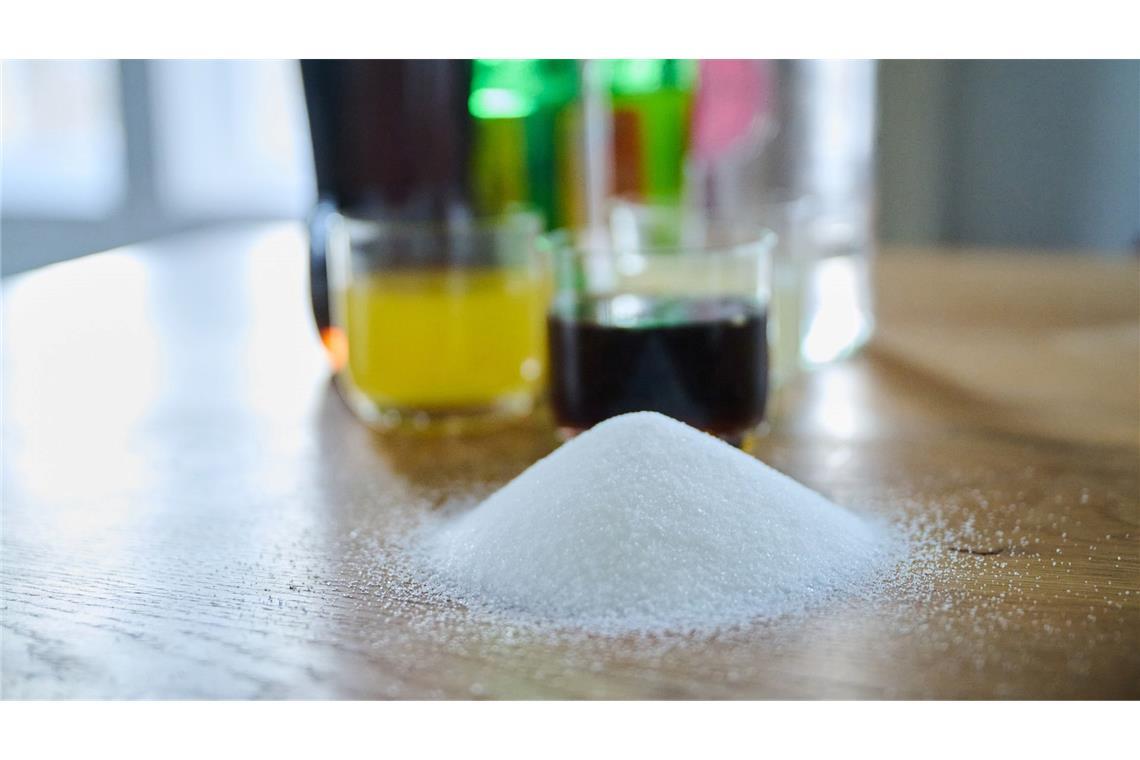 Softdrinks erhöhen Risiko für Diabetes und Herzkrankheiten Drei unterschiedliche Arten Softdrinks und die Flaschen dazu stehen auf einem Tisch, während ein Berg von Zucker davor aufgeschüttet ist