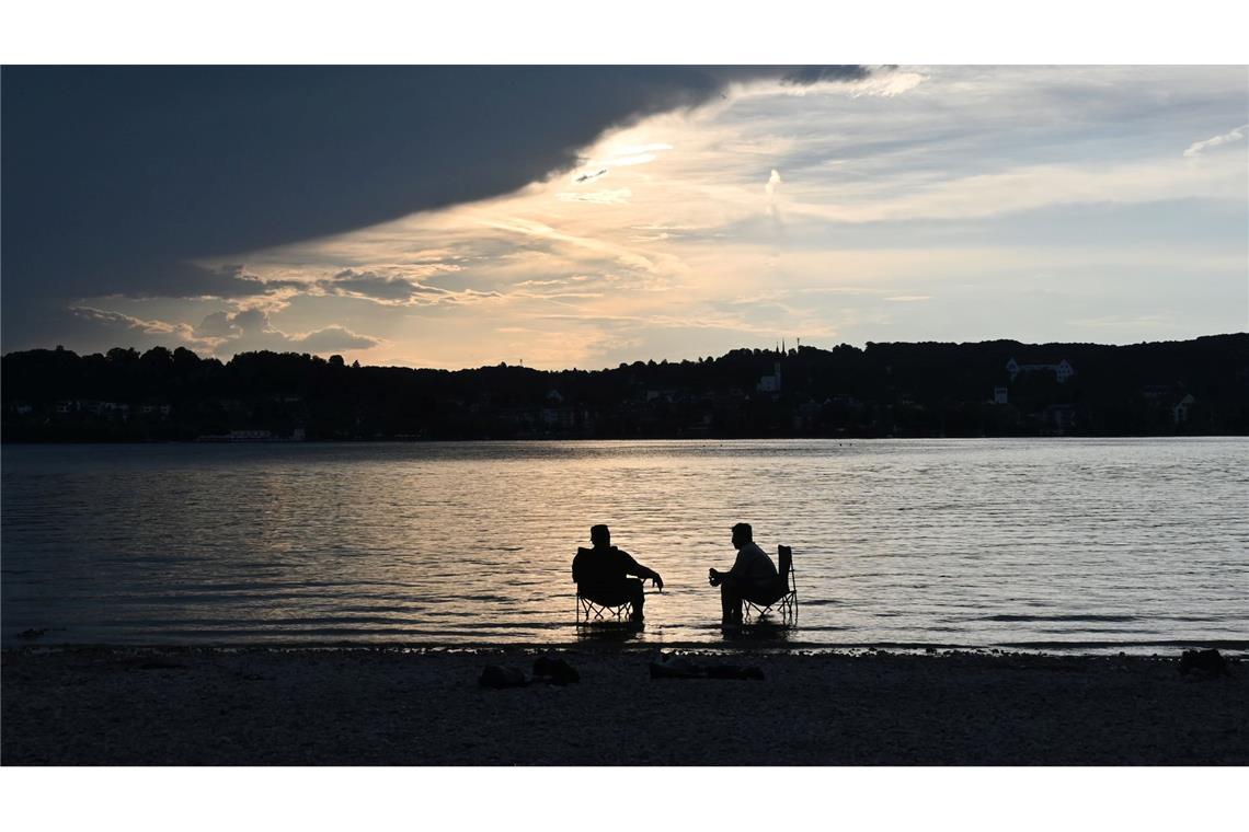Dunkle Wolken ziehen über den Starnberger See während zwei Männer mit Campingstühlen im Wasser sitzen.