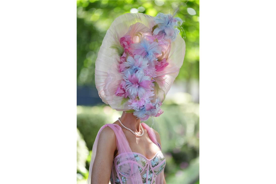 Eine Besucherin von Royal Ascot trägt einen pastellfarbenen Blütenhut in Rosa, Hellblau und Creme, der das Gesicht fast vollständig verdeckt. Das florale Motiv setzt sich im zarten Kleid mit Tüllträgern und Stickereien fort.
