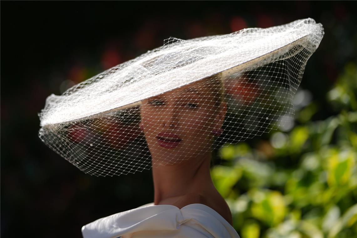 Eine Besucherin von Royal Ascot trägt einen eleganten weißen Tellerhut mit feinem Netzschleier, der das Gesicht teilweise bedeckt. Der reduzierte Look wirkt deutlich klassischer und zurückhaltender als viele der farbenfrohen Statement-Hüte.