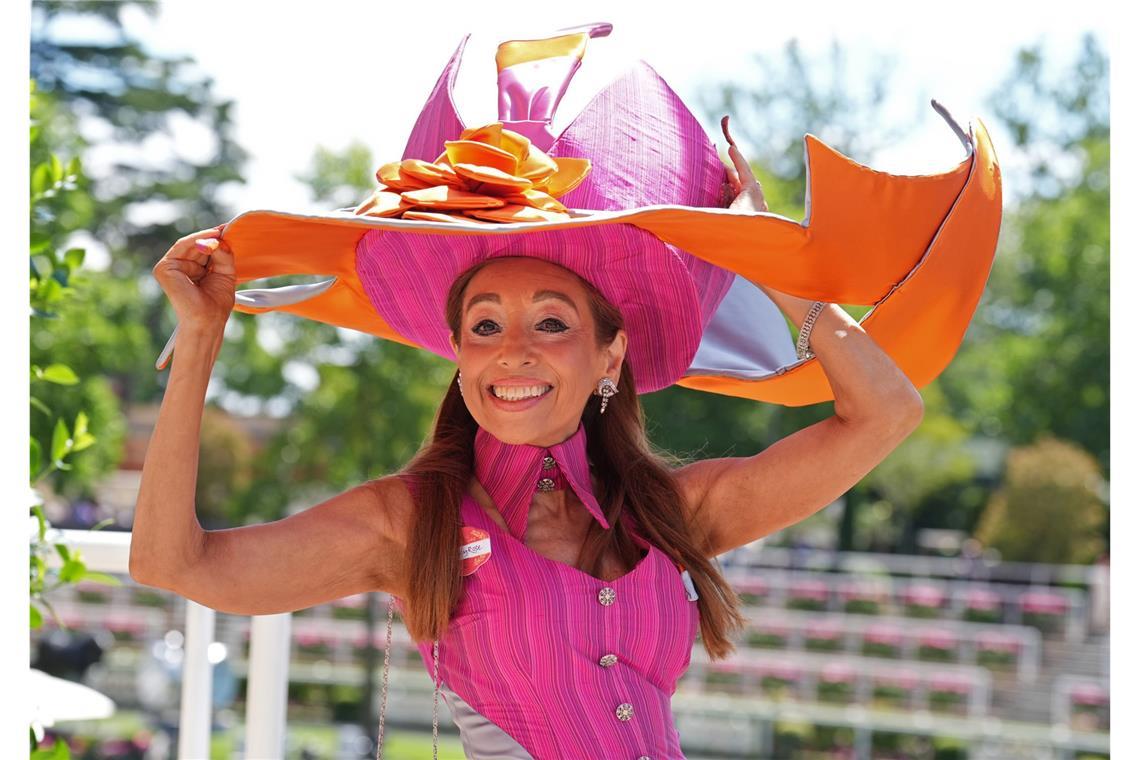 Eine Besucherin von Royal Ascot trägt einen extravaganten Hut in Pink und Orange mit weit ausladender, geschwungener Krempe und Blütendetail. Das knallige Pink des Kopfschmucks wird im ärmellosen Kleid mit hohem Kragen wieder aufgegriffen.
