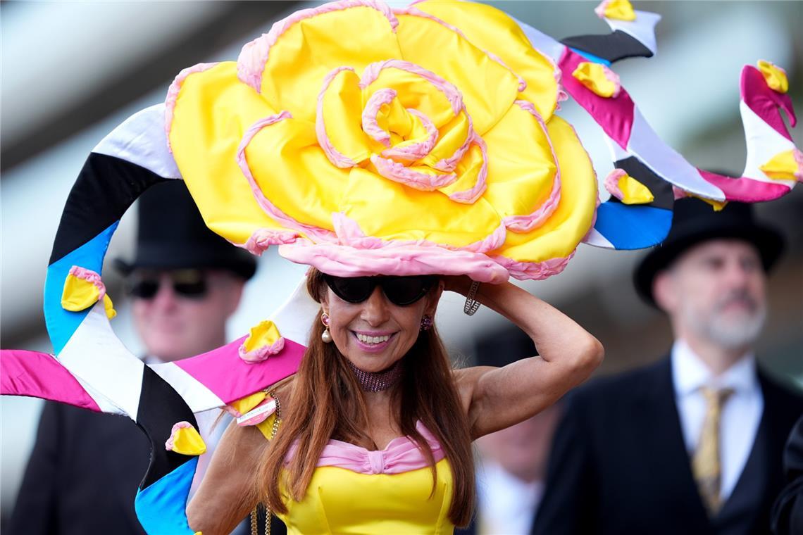 Eine Besucherin von Royal Ascot trägt einen riesigen gelben Blütenhut mit rosafarbenen Akzenten und weit geschwungenen, mehrfarbigen Elementen. Das gelb-pinke Outfit greift die Farben des Kopfschmucks konsequent auf.