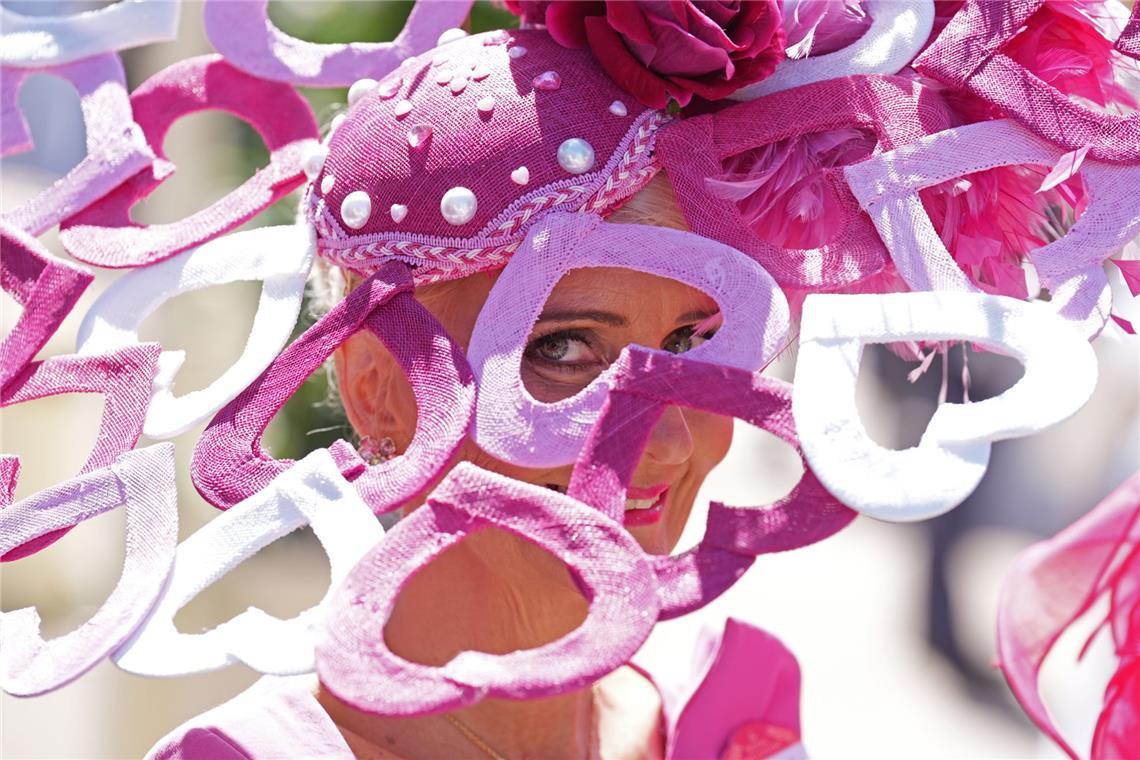 Eine Besucherin von Royal Ascot trägt einen rosafarbenen Hut mit Perlen, Blüte und Federn. Besonders auffällig sind die ausladenden, geschwungenen Elemente in Pink, Rosa und Weiß, die den Kopfschmuck fast wie eine textile Skulptur wirken lassen.