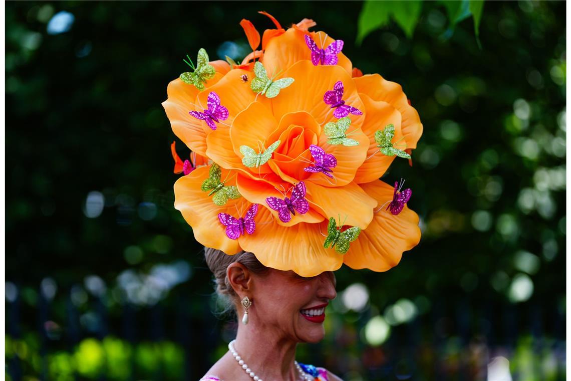 Eine Besucherin von Royal Ascot trägt einen übergroßen orangefarbenen Blütenhut, der mit violetten und grünen Schmetterlingen verziert ist. Der florale Kopfschmuck wird mit Perlenkette, Tropfenohrringen und einem farbenfrohen Kleid kombiniert.
