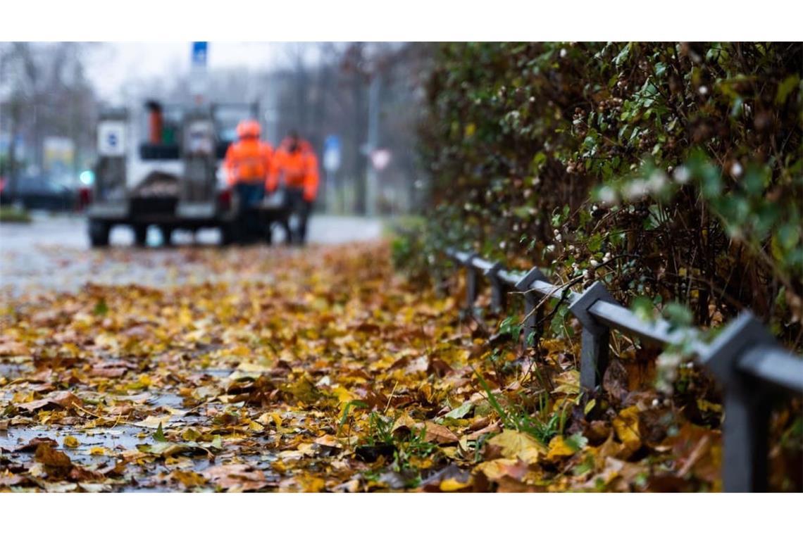 Herbstliches Laub liegt auf dem verregneten Bürgersteig, während im Hintergrund zwei Arbeiter ihre Schubkarre beladen. 