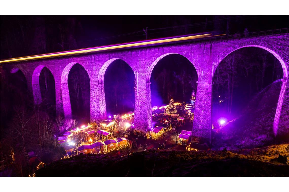 Menschen stehen auf dem Weihnachtsmarkt in der Ravennaschlucht, während über ihnen ein Zug über das Eisenbahn-Viadukt fährt.