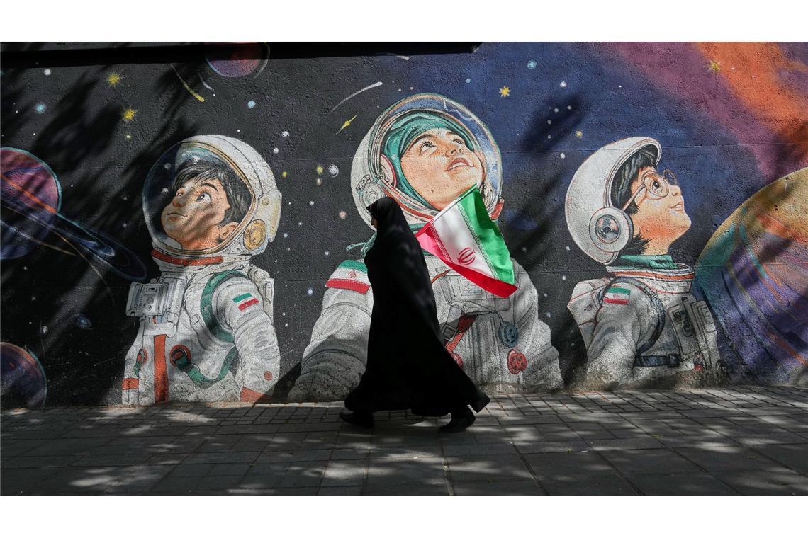 Mit einer iranischen Flagge in der Hand geht eine Frau an einer Wand vorbei, auf der Kinder in Raumanzügen zu sehen sind.