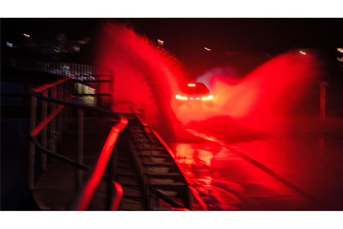 Wasser spritzt, während ein Auto über eine überschwemmte Straße zwischen Gleusdorf (Landkreis Haßberge, Unterfranken) und Busendorf (Landkreis Bamberg, Oberfranken) fährt.