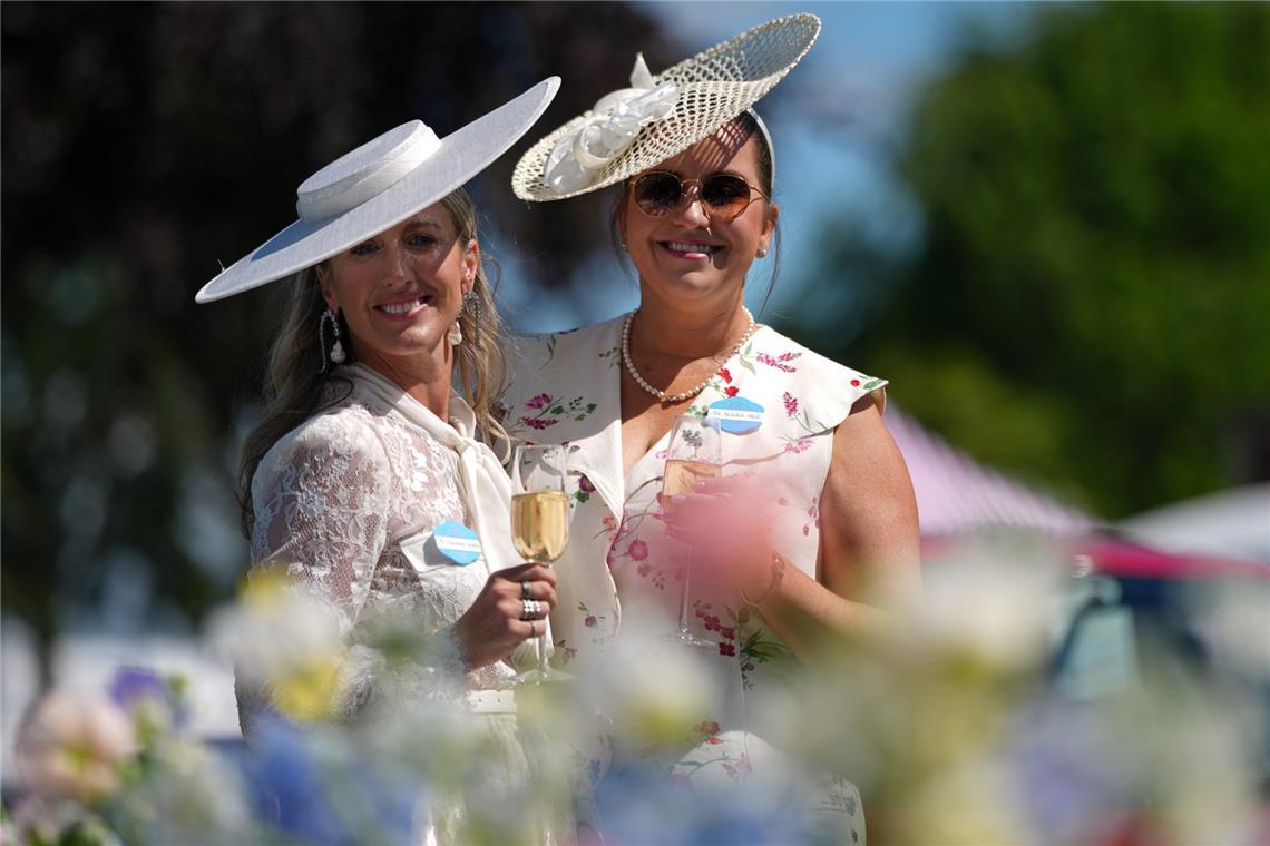 Yallulah Hall und Francesca Gamble (l) zeigen zurückhaltendere Sommerlooks in hellen Tönen: ein weißer Tellerhut zur Spitzenbluse und ein cremefarbener Schräghut mit Blütendetail zum floralen Kleid. Perlen und Champagnerglas unterstreichen den klassischen Ascot-Stil.