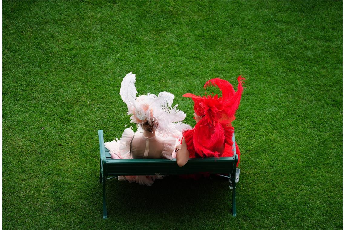 Zwei Besucherinnen von Royal Ascot sitzen in farblich abgestimmten Feder-Looks auf einer Bank: links ein zarter roséfarbener Kopfschmuck mit großen Federn und Tüll, rechts ein dramatischer roter Hut mit ausladenden Federn und passendem Kleid.