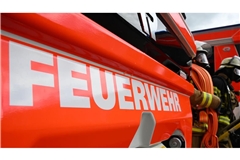 Zwei Feuerwehrleute gehen mit ihrer Ausrüstung zur Brandbekämpfung an einem Einsatzfahrzeug vorbei.